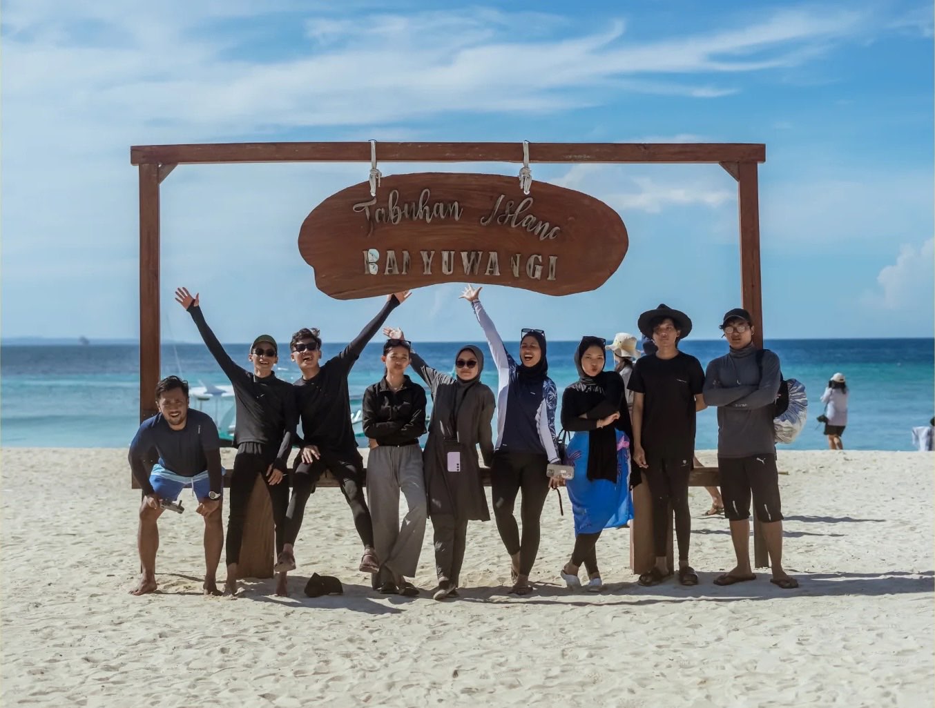 Sebelas orang tim studio by KAMI berpose di bawah papan kayu bertuliskan Tabuhan Island Banyuwangi, di pantai berpasir putih